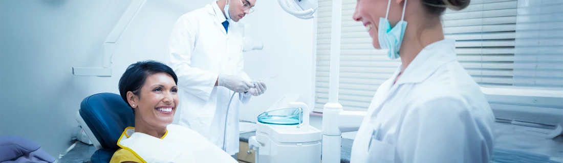 Dental treatment room with a seated patient, two staff members, equipment, and window blinds.